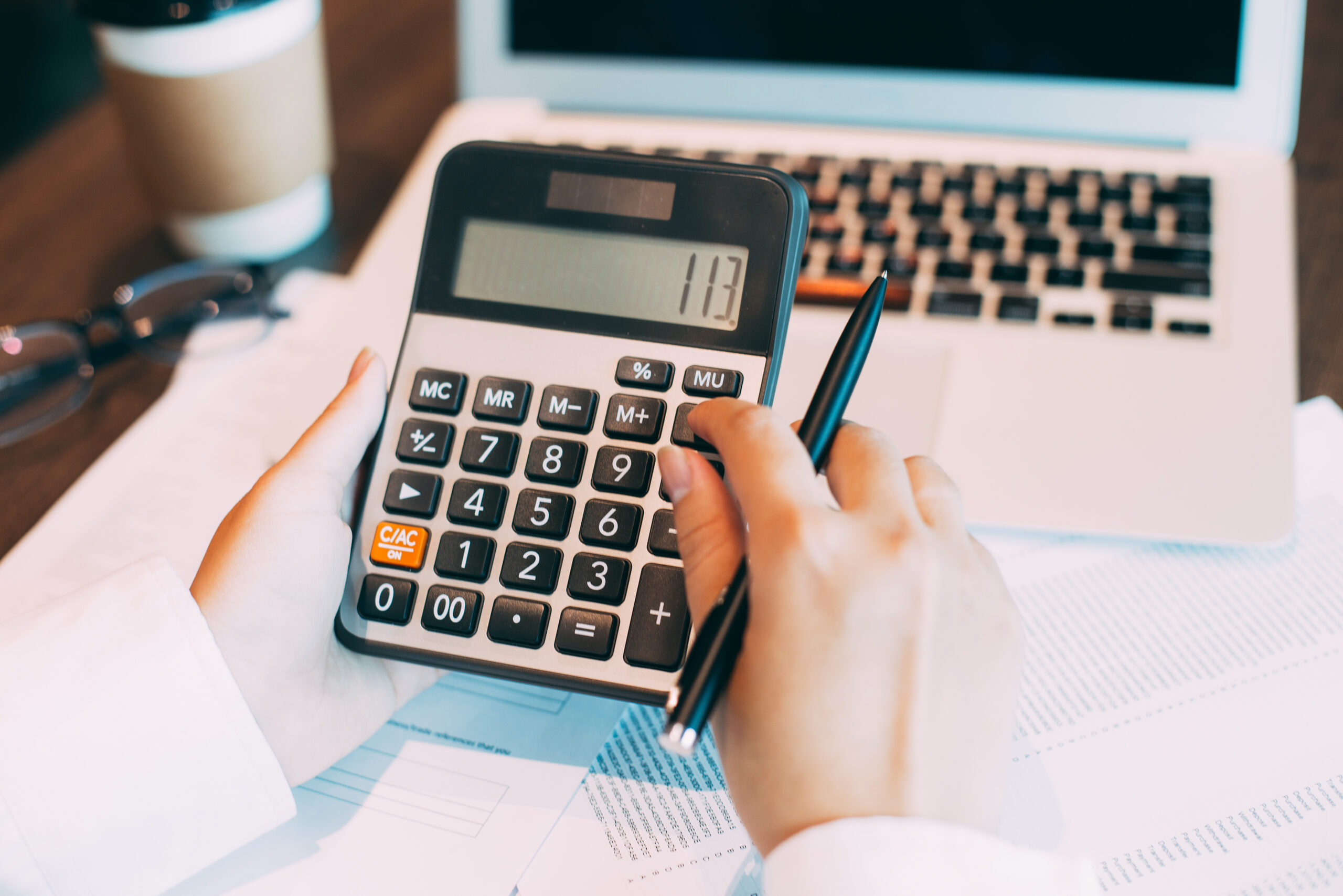 Female accountant sitting at table with laptop, papers, coffee cup and glasses, holding ballpoint pen and using calculator